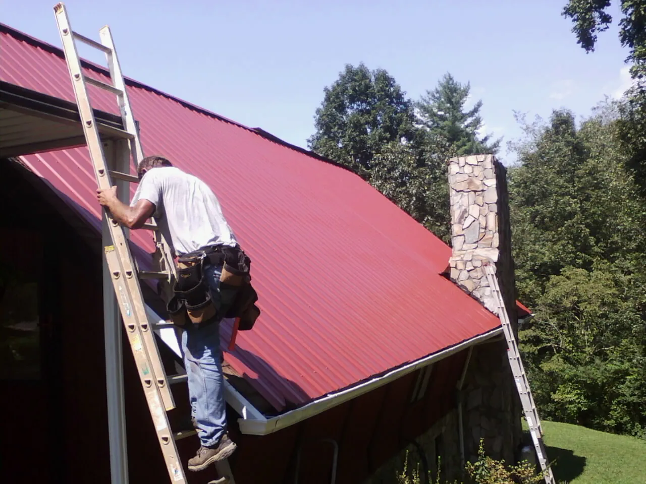 Roofer on a ladder beside a red metal roof and stone chimney — Soffit & Fascia Repair by Roofing Brothers in Dorchester Center